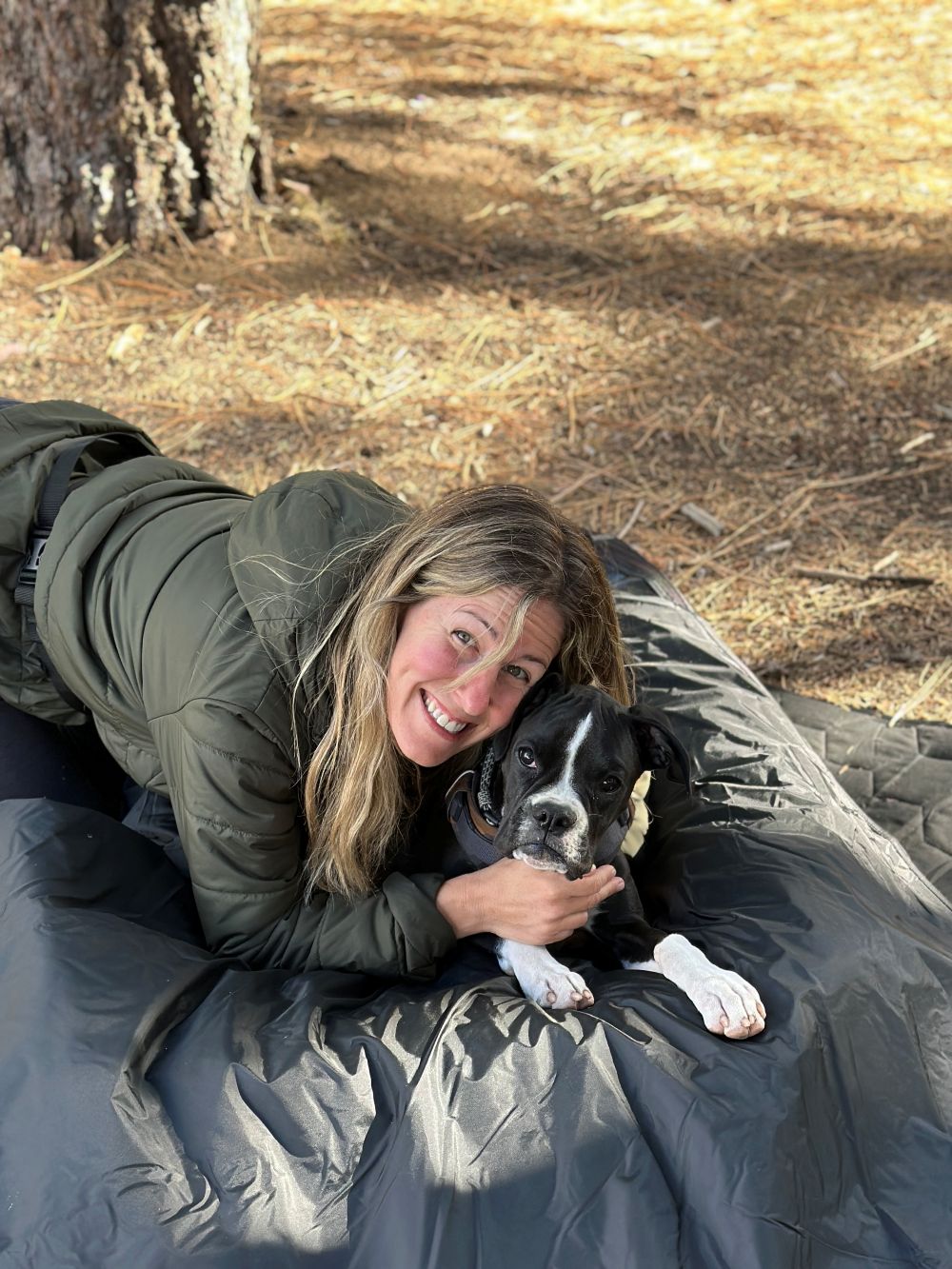 Jenny in outdoor gear cuddling with her dog Bliss on a blanket under a tree during a camping trip.