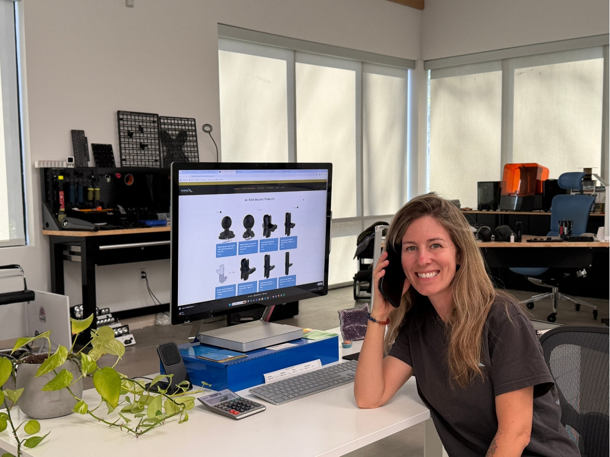 Terra X co-founder Jenny Baumann smiling on the phone at her desk in the Vista, California workshop, with Terra X RAM mount products on the monitor and 3D printers in the background