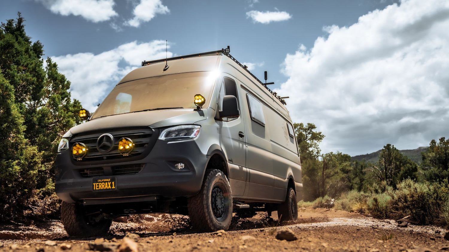 A custom Mercedes Sprinter camper van equipped with heavy-duty Terra X gear, driving off-road on a dusty trail with a custom 